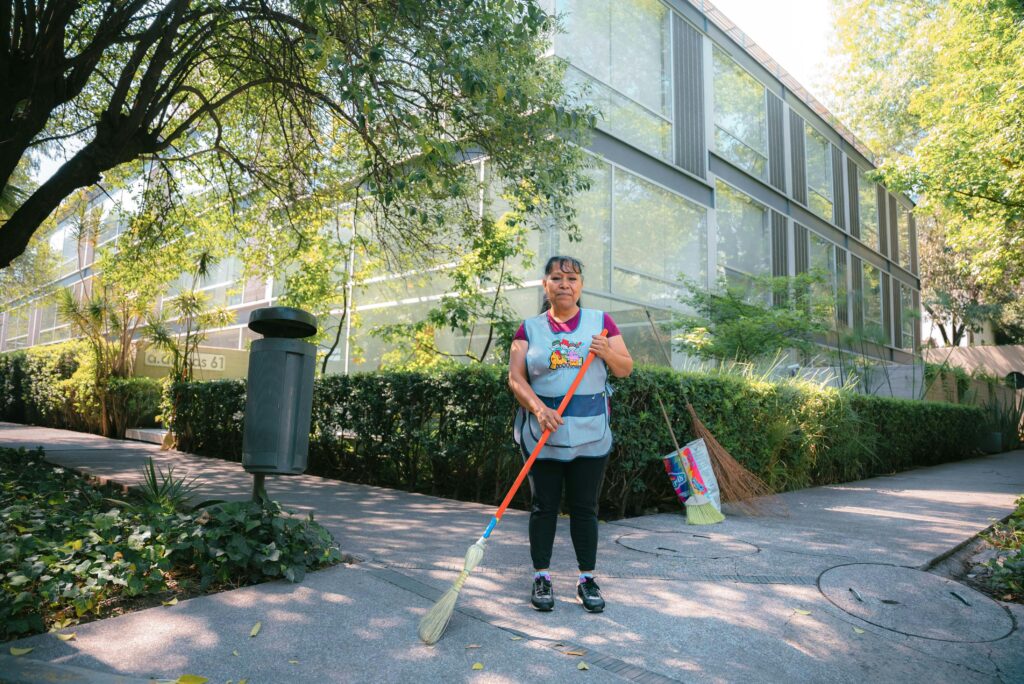 pexels photo 16833890 16833890 A woman casually sweeping a park walkway in Mexico City, surrounded by greenery and modern buildings.